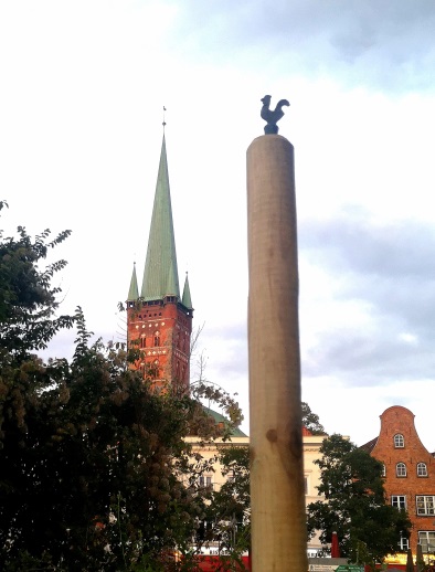 Ein Gusseisener Hahn trohnt auf einem Holzstab. Fotografie aus einer niedrigen Perspektive, sodass der Hahn im Himmel zu schweben scheint.Im Hingergrund der Turm von St. Petri