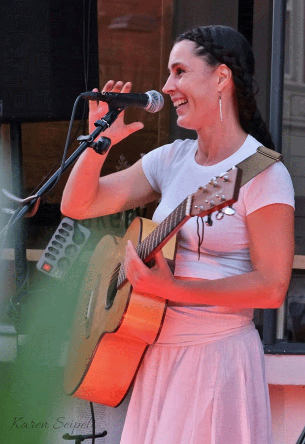 Eine Frau singt ins Mikrofon und hält eine Gitarre in der Hand, sie trägt ein weißes T-Shirt und weißen Rock und hat geflochtene dunkle Haare