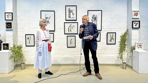 Das Foto zeigt die Eröffnugnsrede auf der Vernissage zur Ausstellung "Ton der Stille" von Lena Römer. Herr Fischer spricht ins Mikrofon, Frau Römer steht vor ihren Kunstwerken.ommensrede auf der Vernissage der Ausstellung "Ton der Stil